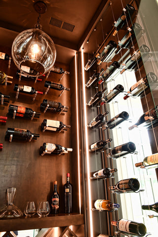 Three glass and cable wine racks mounted in front of a glass window which serves as one wall of a small wooden wine cellar