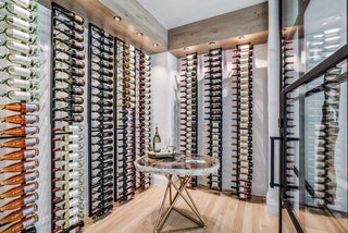 Several columns of floor to ceiling black metal wine racks arranged in a wine cellar with white walls, wooden floors, and a glass door. A table with a wine bottle sits in the centre of the room.