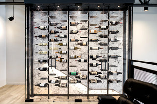 Metal wine racks inside a black-trimmed glass wine wall with a staircase behind.