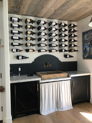Six rows of label-facing wine bottles mounted against a cream coloured wall above a farmhouse sink with a black backsplash.