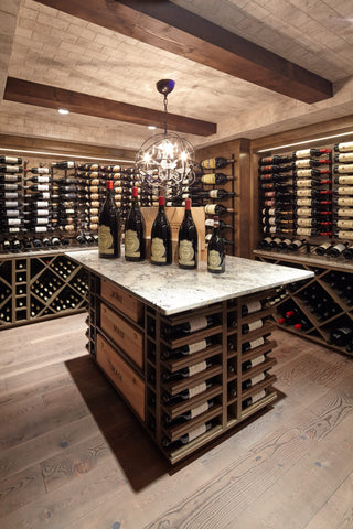 Wine bottles of various sizes arranged largest to smallest atop an island countertop surrounded by walls covered in wine racks. 