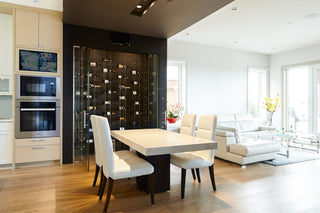 White dining table in front of a black walled windowed wine cellar with a glass door. Inside glass and cable wine racks contain several bottles of wine.